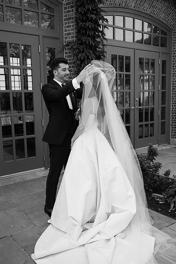 First look moment as groom lifting veil from bride in a strapless gown, bow tie visible, beside ivy-covered brick wall and glass doors