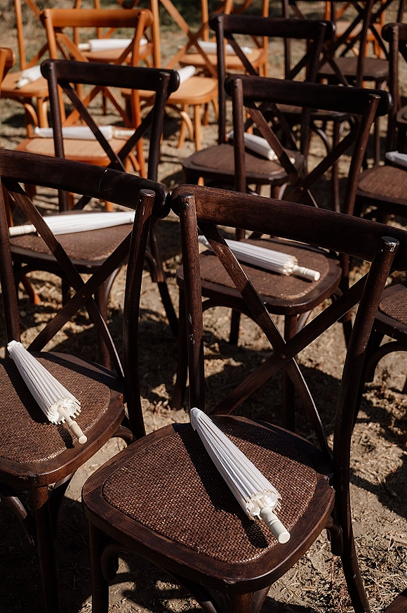 Ceremony seating with cross back ceremony chairs in sunlit outdoor rows, each chair topped with a white paper parasol for guests
