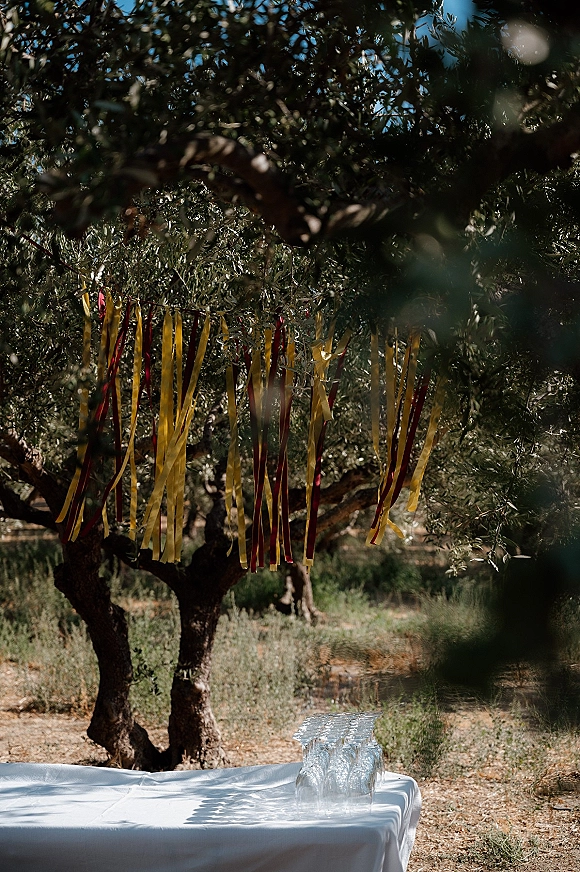 Wedding ribbon decor with a hanging ribbon backdrop draped from tree branches above a white linen table with glassware outdoors
