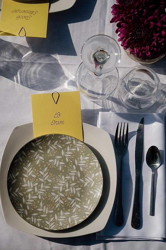 Wedding place setting with a wedding table place card in yellow clipped above patterned plate, white napkin, flatware, glasses, and purple blooms