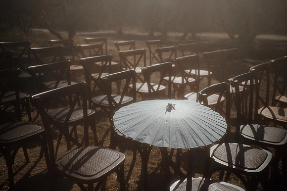 Ceremony seating with outdoor ceremony chairs in neat rows of wooden crossback chairs on a sunny lawn, with a white paper parasol nearby