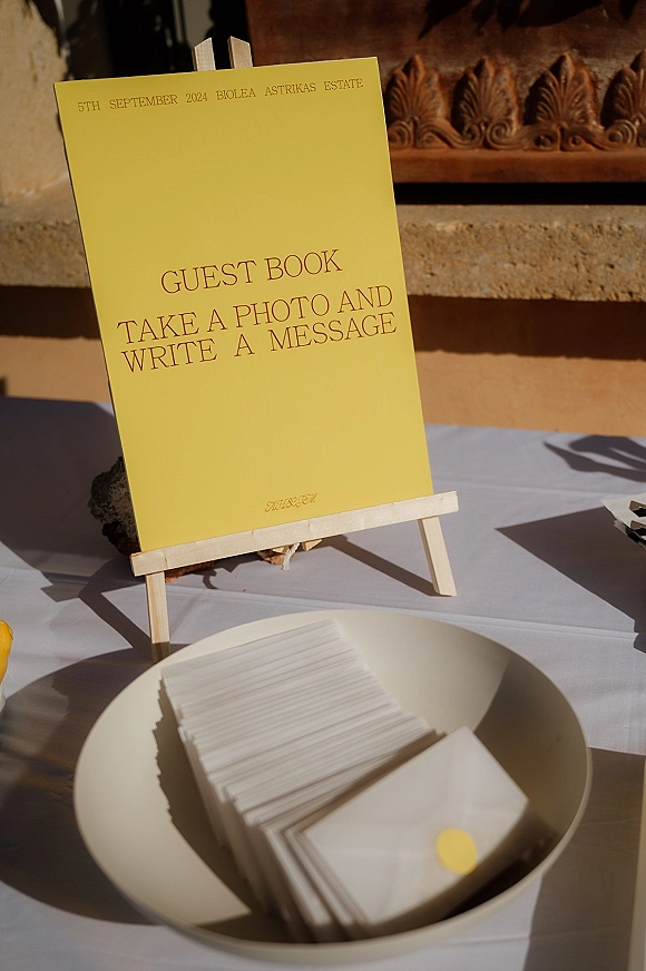 Wedding guest book display with yellow sign on a wooden easel, cards and envelopes in a white bowl on linen table by a stone wall