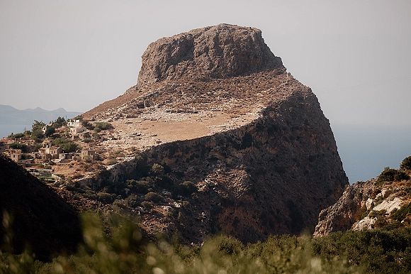 Mountain landscape with a dirt road winding past stone buildings on a rocky hillside, overlooking the sea and distant hazy peaks