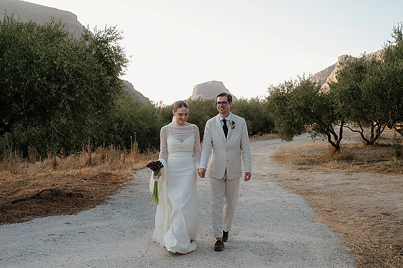 Couple portrait of bride and groom walking hand in hand, bride holding calla lily bouquet on a dirt road through olive trees with mountains behind