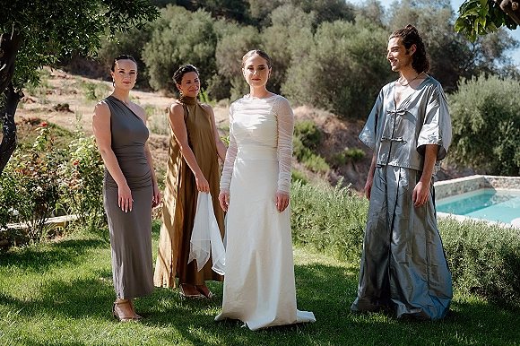 Bridal party portrait with bride in sheer long-sleeve gown and bridesmaids in satin neutral dresses on a garden lawn by a pool