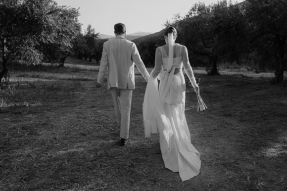 Couple portrait of bride and groom holding hands walking away, her veil and bouquet trailing in an olive grove with mountains beyond