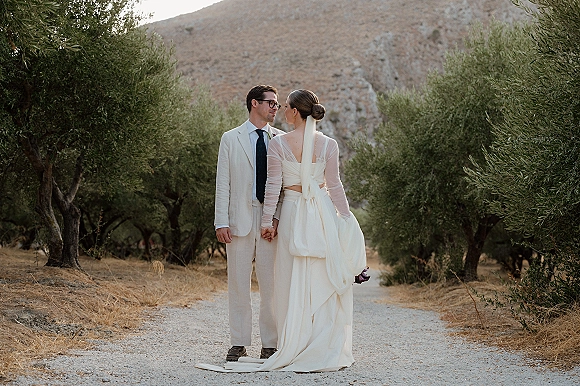 Couple portrait of bride and groom holding hands, looking at each other on a gravel path in an olive grove, her long veil flowing.