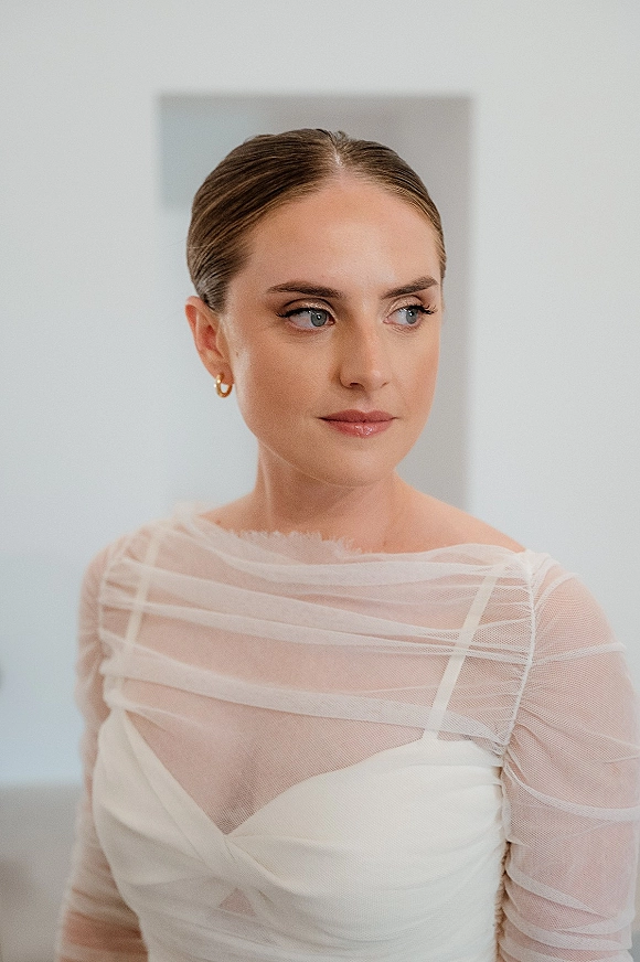 Bridal portrait with classic bridal makeup, bride looking away in a sheer long sleeve gown with gold hoop earrings by a neutral wall with framed artwork