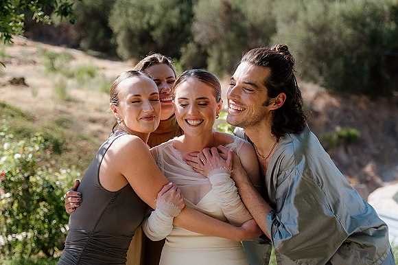 Wedding party portrait of the bride with bridesmaids hugging and laughing, her sheer sleeve dress glowing in sunlit hillside greenery