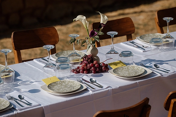 Reception tablescape with an outdoor wedding table setting on a white tablecloth, calla lilies, grapes, patterned plates, and wooden chairs in a dry field