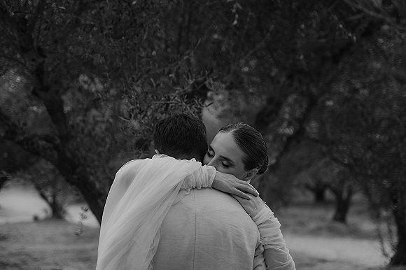 Wedding couple embrace as bride hugs groom with veil draped over his shoulder, eyes closed, in a quiet tree grove outdoors