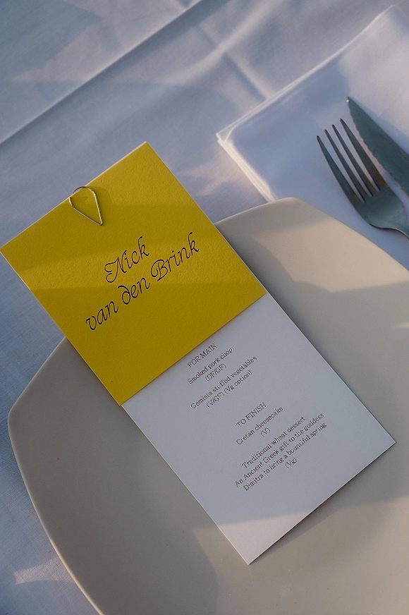 Wedding place card in yellow clipped with a silver paperclip atop a printed menu, set on a white plate with fork and knife in sunlit shadows