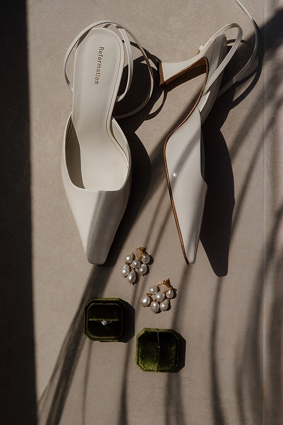 Bridal shoes flatlay with white slingback heels, pearl cluster earrings, engagement ring and velvet boxes on neutral stone in window light shadows