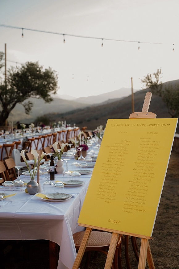 Wedding seating chart on easel with a yellow board beside long banquet tables, calla lilies, and string lights at a mountain-view reception