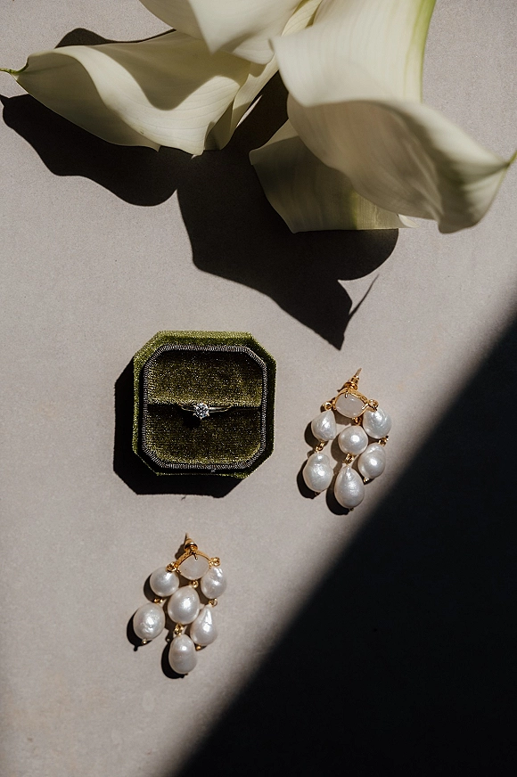 Engagement ring beside a diamond solitaire ring in a velvet ring box with pearl drop earrings and calla lily on a neutral surface in hard sunlight shadows