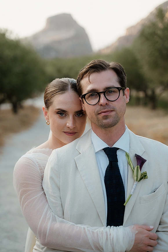 Couple portrait of bride hugging groom from behind, her sheer-sleeved gown against his cream suit with calla lily boutonniere by mountain road