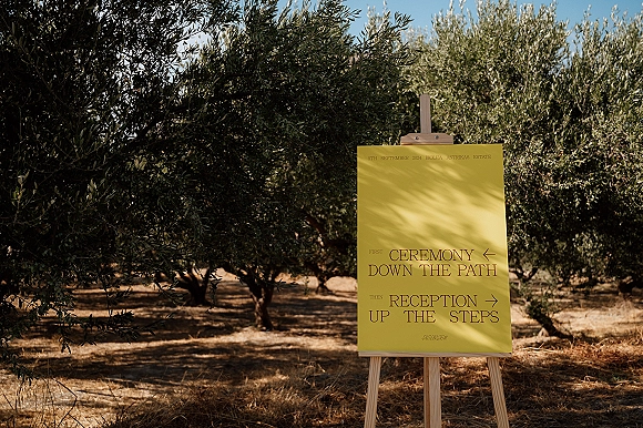 Wedding signage with a wedding direction sign on an easel in an olive orchard, dry grass under blue sky with sunlight shadows
