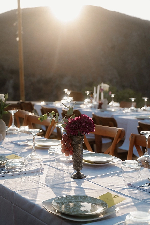 Reception tablescape with an outdoor reception table set in white linen, burgundy dahlia and grapes, wooden chairs, and mountain sunset backdrop