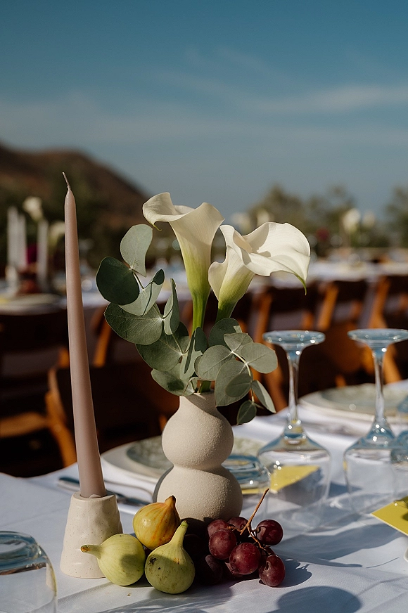 Reception tablescape with an outdoor wedding tablescape look, white calla lilies in a ceramic bud vase and taper candles on long tables under blue sky