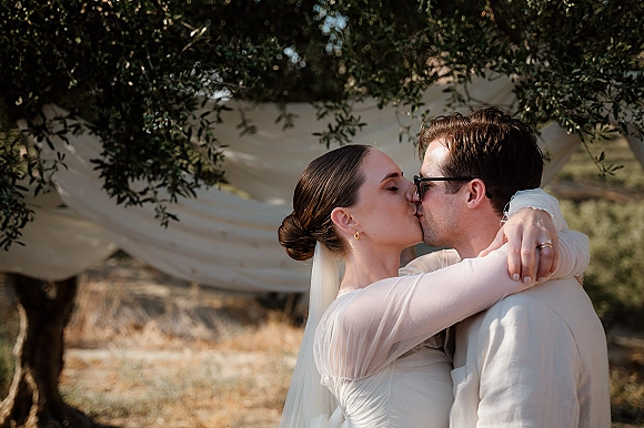 Wedding kiss portrait of bride and groom kissing, her veil flowing as he wears sunglasses, framed by draped fabric and tree branches