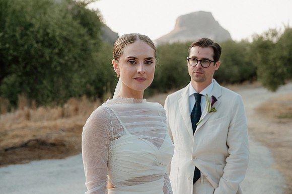 Couple portrait in an outdoor wedding portrait on a dirt road, bride in veil and sheer-sleeved dress with groom in white suit, mountains behind