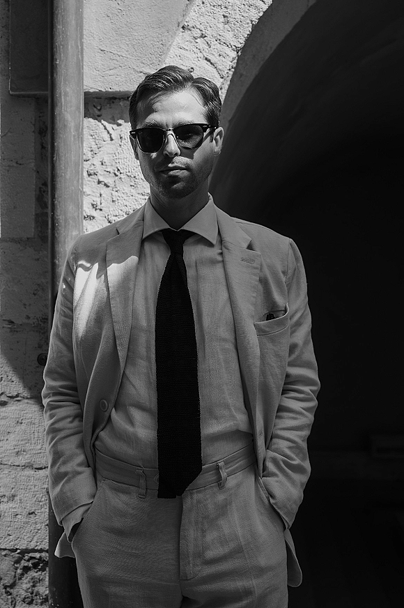 Groom portrait in sunglasses wearing a light suit with dark tie and pocket square, hands in pockets by a stone archway in shadow