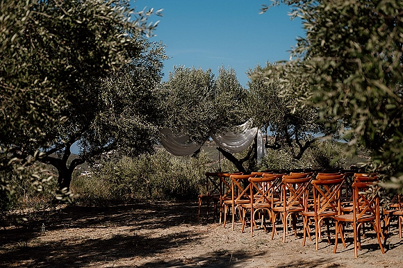 Ceremony setup for an outdoor wedding ceremony with wood chairs in aisle rows and fabric draping beneath olive trees on a hillside