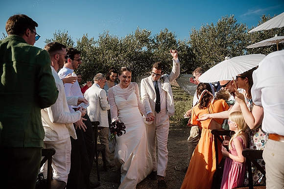 Wedding recessional as bride and groom walk the aisle, bouquet in hand, guests cheer under umbrellas amid confetti in a sunny field ceremony