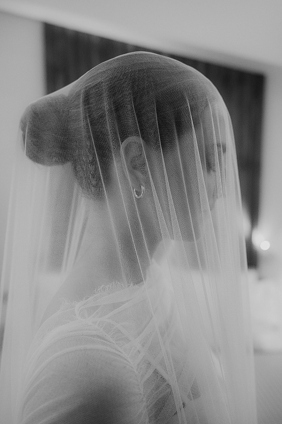 Bridal portrait of a bride under veil with tulle softly covering her face, hoop earrings and gown lit by window light in an indoor room