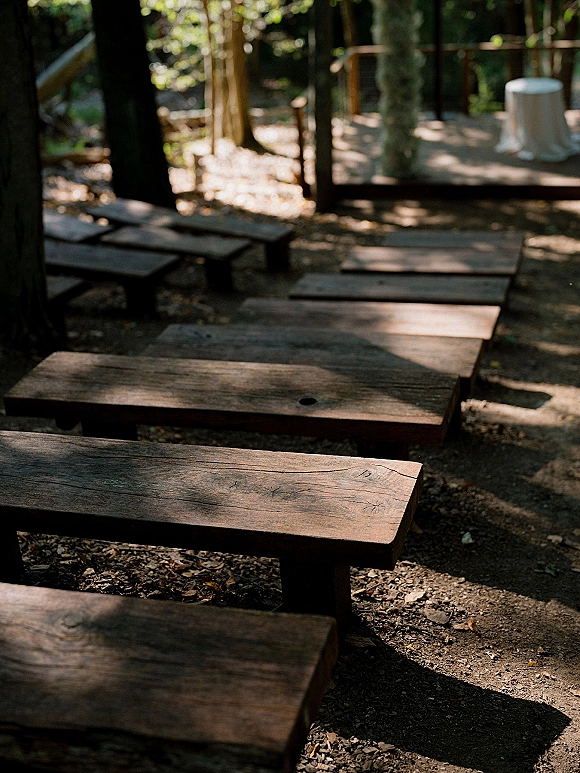 Ceremony seating with rustic wedding benches lined along a dirt aisle, wood benches on a deck beneath sunlit forest trees