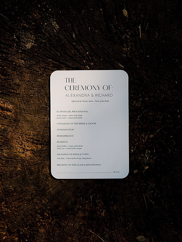 Wedding ceremony program with a printed program card in black and white, arranged on a tree stump showing rustic wood grain texture
