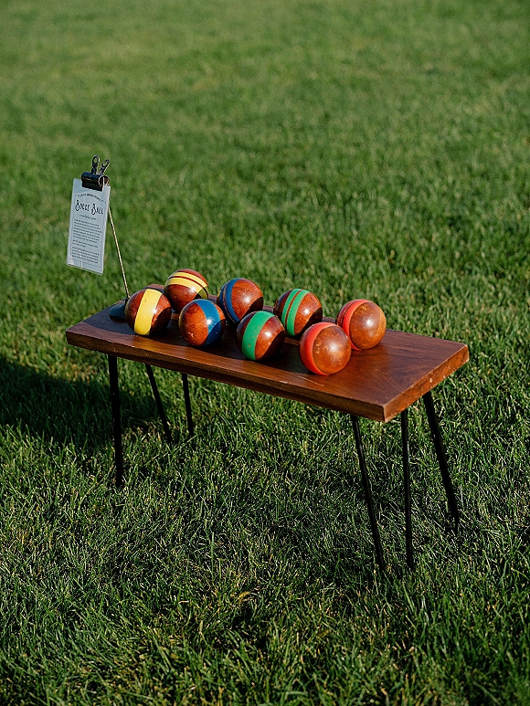 Wedding lawn games setup with colorful bocce ball wedding set, printed rules sign on a wooden table, and balls on grass lawn