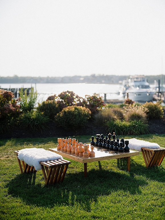 Lawn game setup with a giant chess set beside a low table, wooden stools, and faux fur throws on a waterfront marina lawn