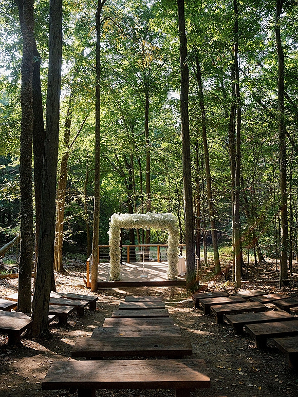 Outdoor ceremony setup with white floral arch on a wooden platform, rustic benches and aisle steps, set in a sunlit forest clearing