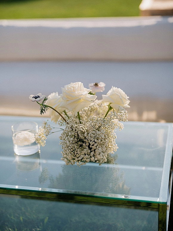 Wedding centerpiece of white roses and baby’s breath with anemone in a glass vase, beside a votive candle on a glass table, lawn seating behind