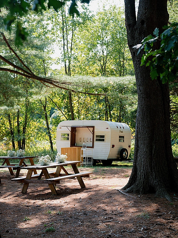 Outdoor reception setup with wood picnic tables, benches, and white floral centerpieces beside a vintage camper drink station in forest shade