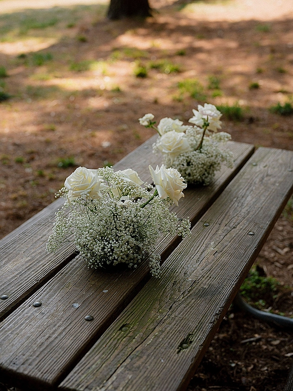Bridesmaid bouquets with white roses and baby’s breath arranged on a rustic wood picnic table in an outdoor park setting