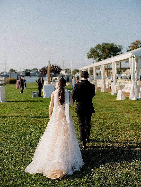 Wedding couple walking across a waterfront lawn, bride in tulle gown with long train and veil beside groom in black suit under tent