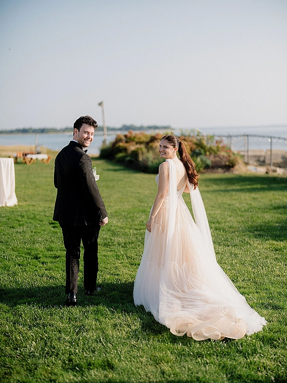 Couple portrait of bride and groom walking away then looking back, her cape veil and long train flowing on a waterfront lawn under clear sky