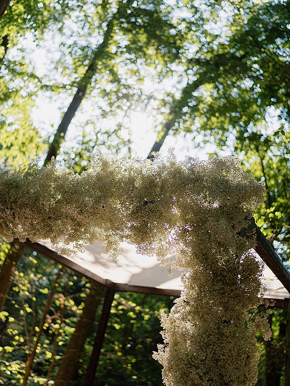 Wedding ceremony arch adorned with white floral installation on a wood frame, set among sunlit forest trees and lush green foliage