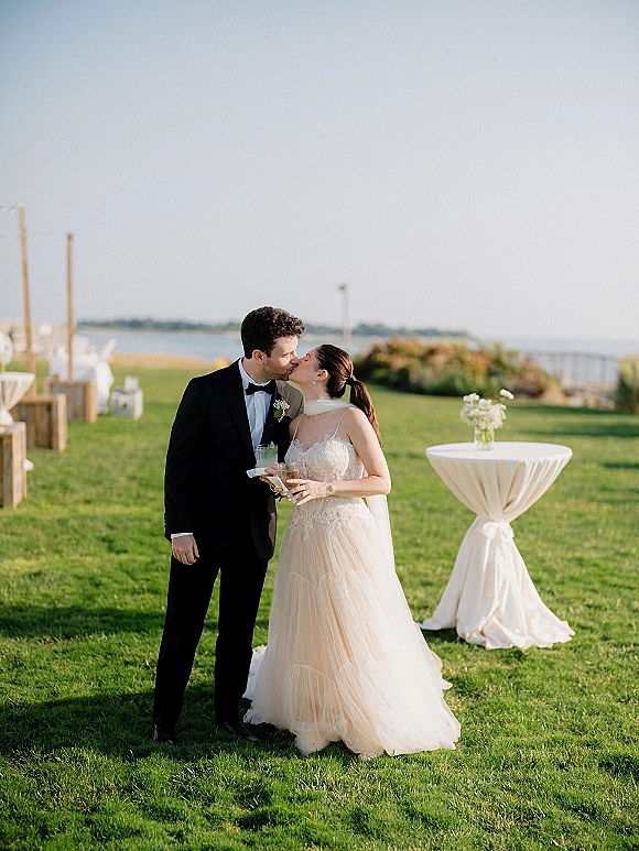 Wedding kiss portrait of bride and groom kissing at waterfront cocktail hour, holding drinks beside a high-top table on an ocean-view lawn