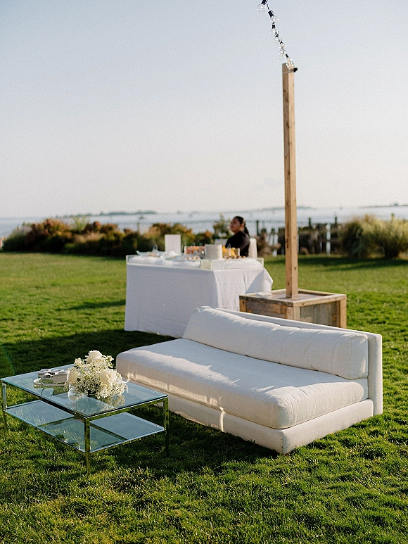 Wedding lounge setup with a white outdoor sofa, glass coffee table, and floral centerpiece on a lawn overlooking the ocean view
