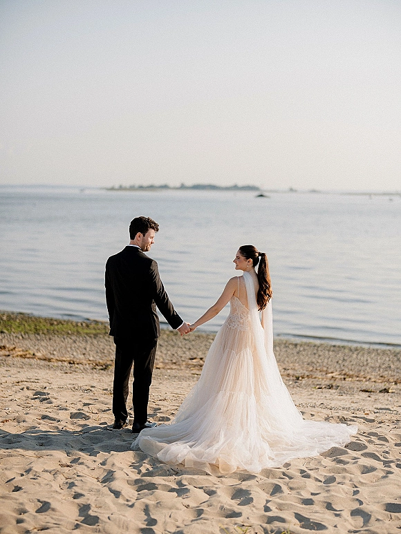 Couple portrait at a beach wedding, bride and groom holding hands on the shoreline with long veil and ocean backdrop, distant islands ahead