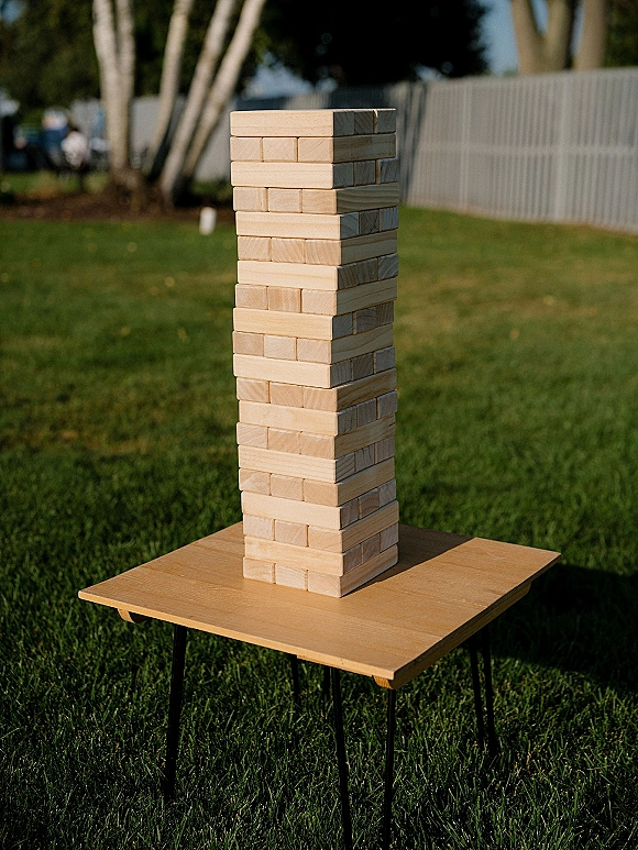 Giant jenga game with stacked wood blocks on a wooden side table on a grass lawn, with trees and fence behind