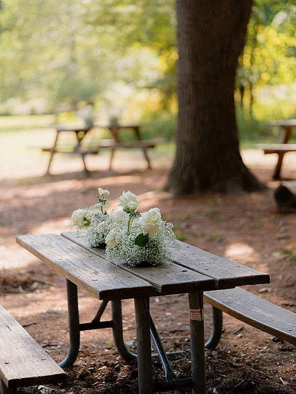 Wedding centerpieces with white rose centerpieces and baby's breath on a wood picnic table in an outdoor park with trees and benches