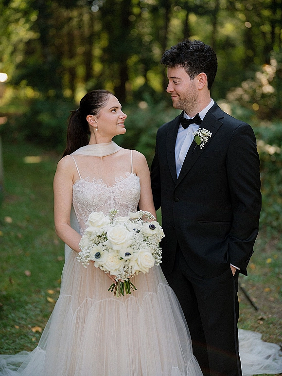 Couple portrait of bride and groom looking at each other, she holds a white rose bouquet with baby's breath in a woodland setting