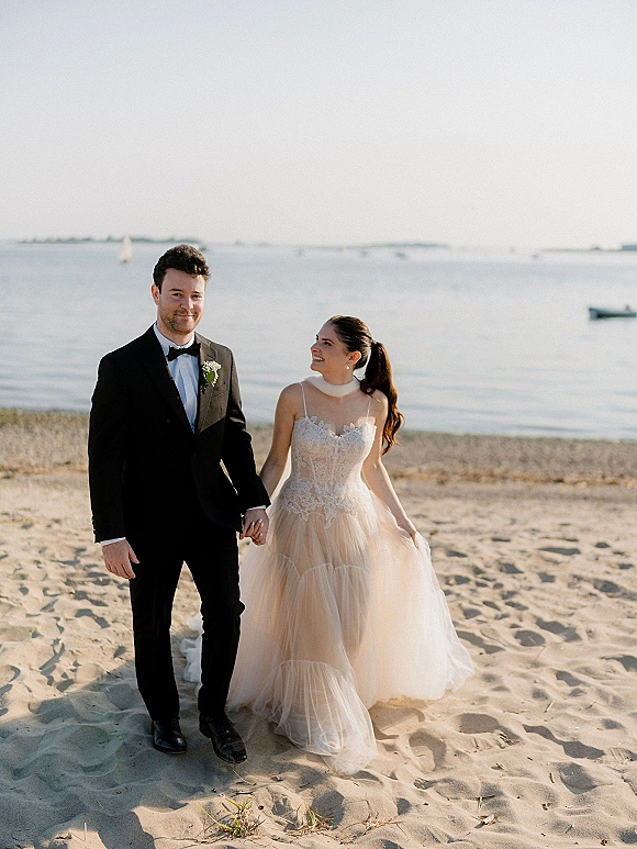 Couple portrait of bride and groom holding hands on a sandy beach, her lace tulle gown and his black tuxedo with boutonniere by the ocean