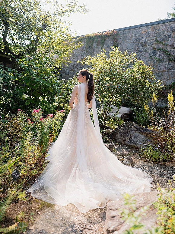 Bridal portrait of a bride from behind in a backless wedding dress with long veil, tulle skirt, and earrings on a sunlit garden path by a stone wall