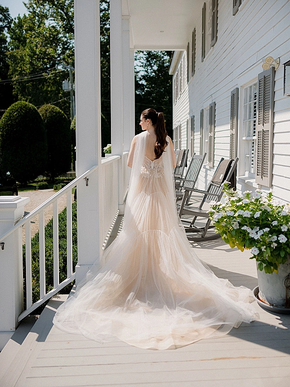 Bridal portrait of a bride from behind in a backless wedding dress with cathedral veil and train, standing on a white porch with rocking chairs