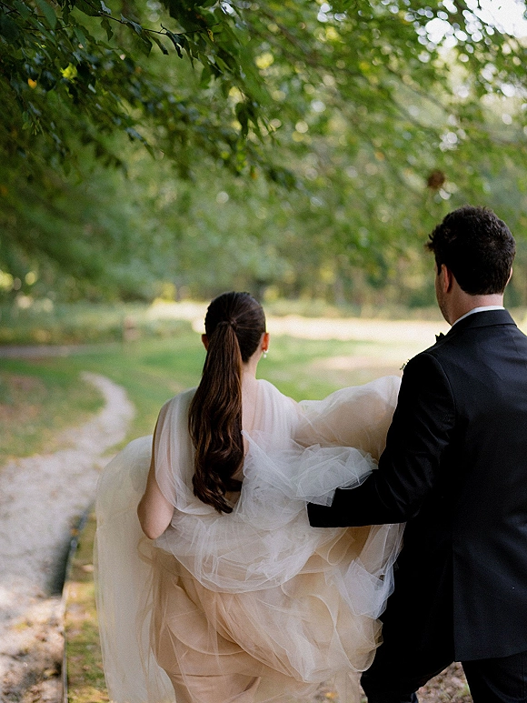 Couple portrait of bride and groom walking away on a park path under a tree canopy, her tulle wedding dress train and his tuxedo visible